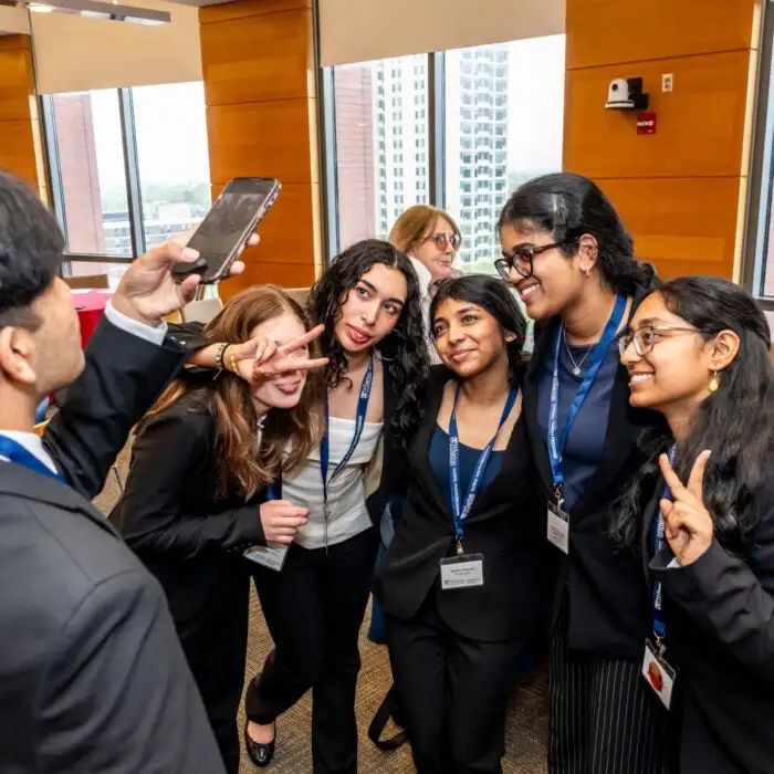 A small group of people in business attire takes a selfie indoors. Some make peace signs, and all appear to be enjoying the moment. They are in a room with large windows and city views.
